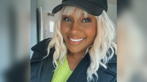 A woman is seen close up. She is smiling at the camera. She is wearing a green jumper under a black jacket and a black cap.