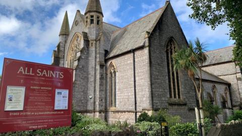 A church building. There is a red sign next to it which says "All Saints". There is a palm tree to the right of the image. The sky is blue with white clouds. 
