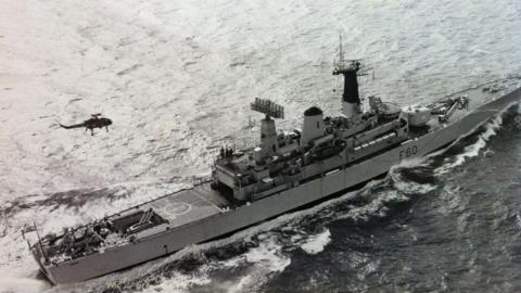 An aerial shot of HMS Jupiter. The black and white image shows a long, slim battleship at sea, with a helicopter flying over it.