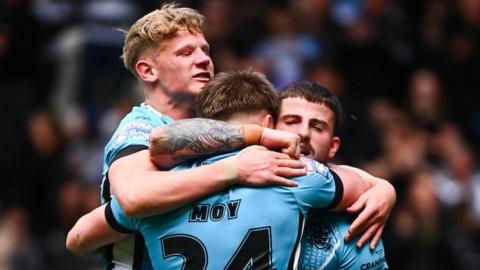 Hull FC players Lloyd Kemp (left) and Amir Bourouh (right) hug try-scorer Logan Moy, all players dressed in light blue.