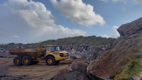 A yellow dumper truck delivering large granite boulders on a sandy beach on a sunny day