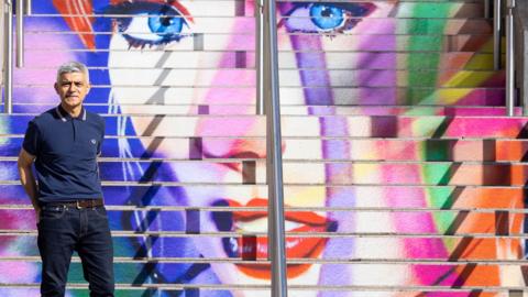 Mayor of London Sadiq Khan, dressed in blue jeans and a blue polo shirt, standing in front of the Swiftie Steps, an outdoor staircase painted with Taylor Swift's face in a graffiti style mural in white, blue, pink, orange, purple and green at Wembley Park, north west London.