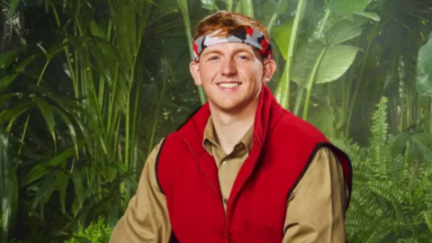 Angry Ginge in a promotional picture for I'm a Celebrity. He wears a red gilet, camo bandana and is smiling.