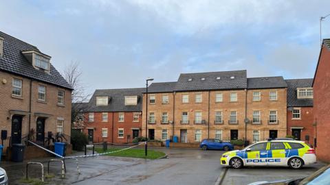 A police car parked in a residential street, opposite a house with police tape blocking off the outside.
