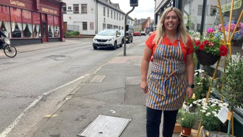 A smiling woman wearing a florist's apron standing outside a flower shop on Devizes Road in Swindon with a view down the road to the left.