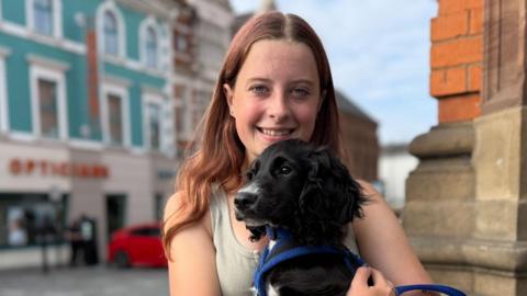 A teenager with long mousy brown hair smiling at the camera. She is wearing a green tank top and standing on the high street. She is holding a dog in her arms. The dog has a blue collar.