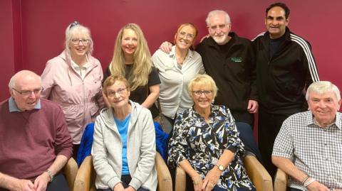 A group of nine people sitting and smiling and looking into the camera. They are smiling. There are two elderly men on the front row and two women. In the back row there are three women and two men. 