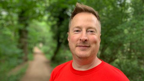 James Houghton is wearing a red t-shirt standing on a path in the forest, surrounded by greenery and trees