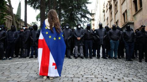 A girl draped in a Georgian and European flag stands facing a dozen men dressed in black