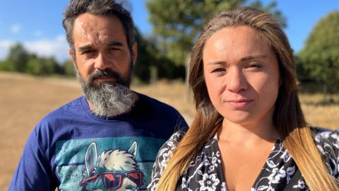 A man and woman sitting one slightly behind the other. The man has a greying beard and is wearing a blue T shirt. The woman has long brown hair wearing a black and white patterned dress 