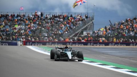 A Formula 1 car on the Silverstone circuit. The car is green, black and silver. In the background is a grandstand full of fans looking on.
