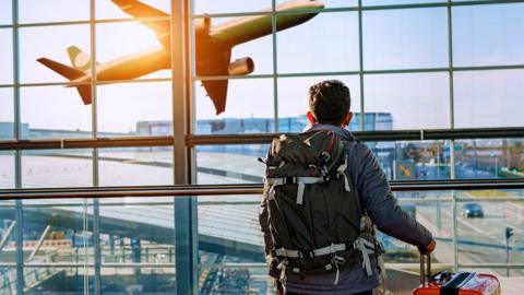 Male tourist is standing in airport and looking at aircraft flight through window. He is holding tickets and suitcase