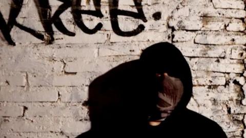 A man standing in shadow and further concealed by a black hoodie and a bandana over his face, in front of the wall of a cartel safe house, which has crumbling brick and fading white paint. There is unintelligible graffiti above his head.