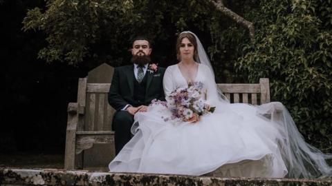 A bride and groom sit side-by-side on a wooden bench looking at the camera, with expressionless faces. The groom is wearing a dark suit with a pink flower attached to his jacket. The bride is wearing a long white dress draped over the side of the bench, and is holding a bouquet of flowers. 