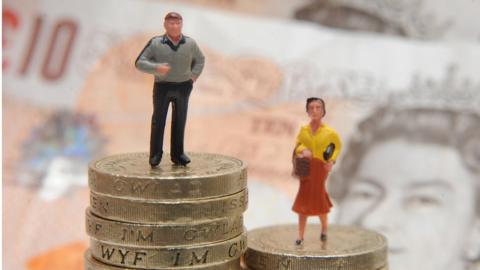 A man standing on a pile of pound coins, and woman standing on a smaller pile of pound coins