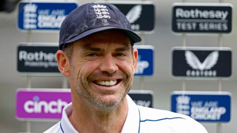 Smiling James Anderson standing in front of a board of sponsers, reading Cinch and We Are England Cricket. He is wearing an England cap.