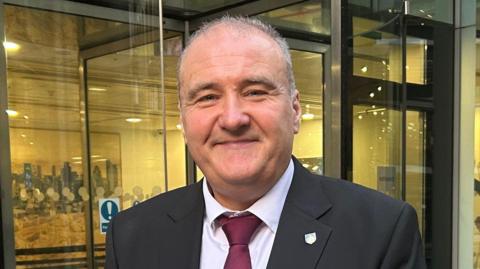 Lee Castleton has receding grey hair and is wearing a black blazer over a white shirt and burgundy tie. He is smiling while standing in front of a glass building.