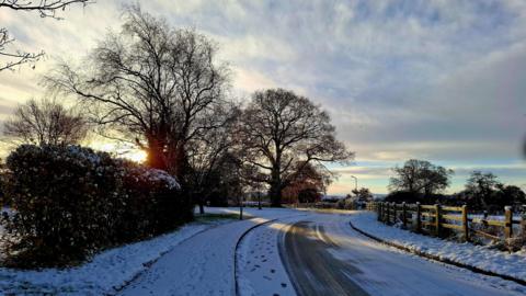 A snowy road with footprints in the snow. There are trees and hedges on the left, with the sun peeking through the branches of a large tree. On the right is a field behind a wooden fence. There are lines in the road where cars have driven.