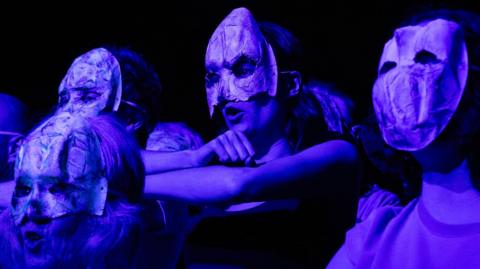 A group of performers in masks and bathed in blue lighting