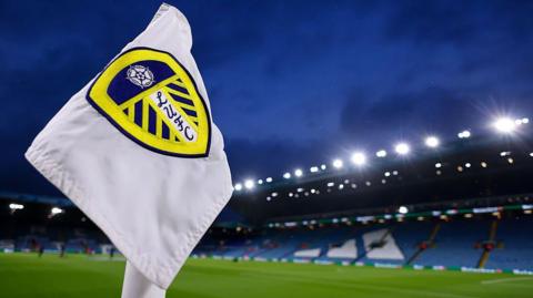 A white corner flag with a Leeds United badge in the middle and Elland Road in the background