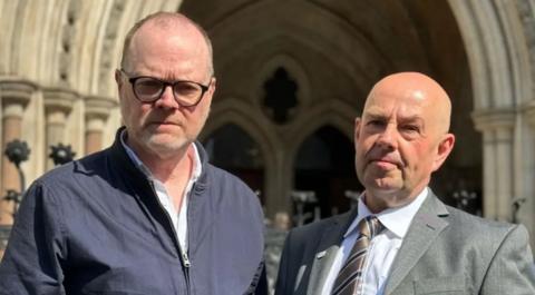 Two men face the camera. One is wearing a dark jacket and white shirt. He has dark rimmed glasses and a grey short beard. The man on the right is wearing a grey suit, white shirt and grey tie. He is balding.