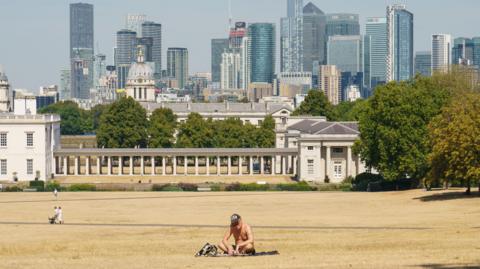 A man sits on a rug on dried out yellowing grass in a London Park in front of a building with the skyline of London in the background