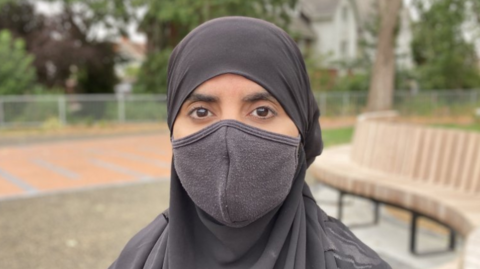 A woman wearing a black hijab and black face mask stands in front of a grassy area of a residential street. 