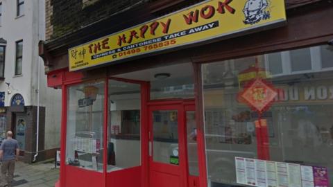 stree4t view of the happy wok with a yellow sign and a red frame work around the takeaway windows