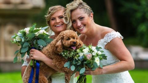 Emma (left) and Helen (right). Both women are blonde and wearing wedding dresses. They are both holding flowers and their dog, which is small with brown curly fur. They are looking directly at the camera and smiling.