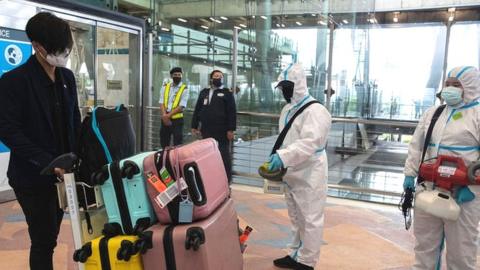 Suvarnabhumi airport staff wearing Personal protective equipment (PPE) suits spray sanitizers on a passenger's luggage on arrival at the airport.