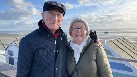 A man and a woman in their 70s smiling at the camera. The man is wearing a black hat and a navy coat. The woman is wearing a khaki coat and a white scarf and hat. Thorpe Bay is behind them