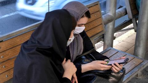Two women look at a mobile phone at a bus stop in Tehran, Iran (30 May 2021)