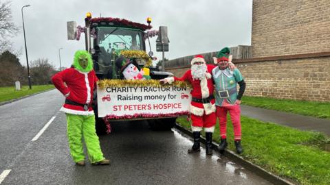 Three men dressed as the Grinch, Father Christmas and an elf are standing near a tractor which says Charity Tractor Run Raising money for St Peter's Hospice