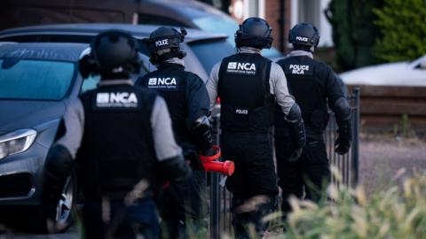NCA officers dressed in helmets, stab vests and other protective clothing walk towards a house. One officer is carrying a red battering ram.