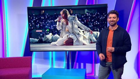 Ricky in Newsround studio with Taylor Swift performing in screen behind him