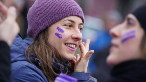 A woman wearing a purple hat puts purple face paint on her cheeks at march to mark International Women's Day