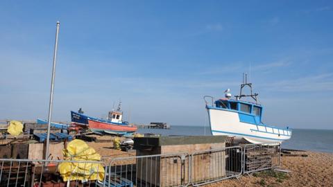 Two boats sit on the beach with blue sea behind and bright blue sky with wispy white clouds above