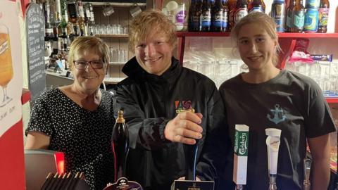 Ed Sheeran behind the bar at The Rumburgh Buck, with landlady Stella Cattermole and another staff member