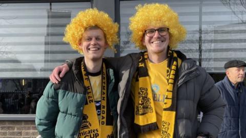 Two male Maidstone United fans wear bright yellow wigs and club kits and smile at the camera