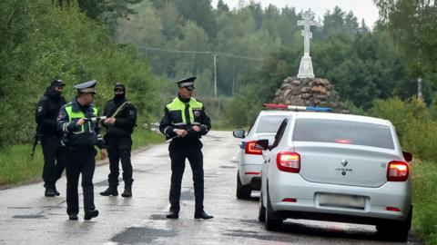 Police officers stand next to a car on a road with trees in the background, as they block the road following the incident