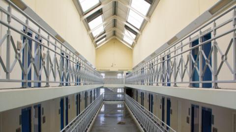Empty corridors in prison with yellow walls and lots of doors open. Building has lots of natural light and is multi-storey. 