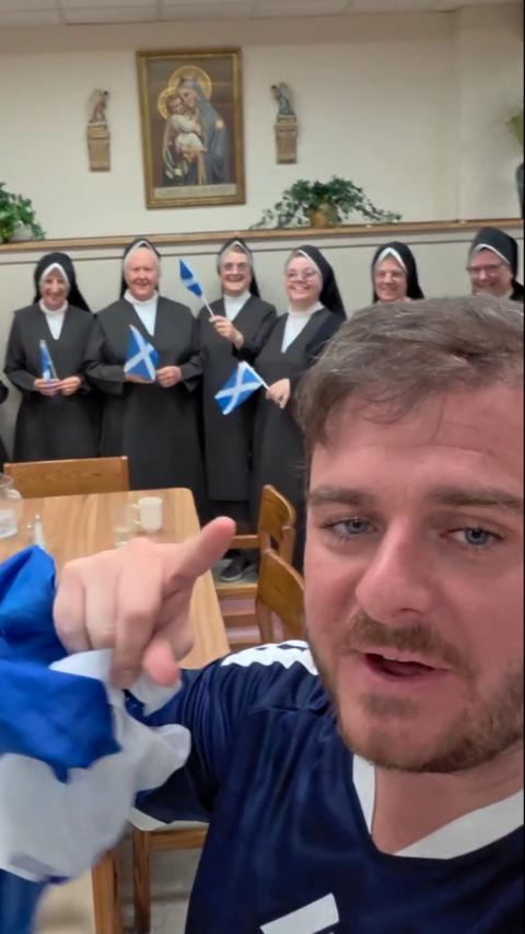 A man with brown hair and blue eyes with a short beard and moustache points at the camera. He is holding a Scotland flag and wearing a blue and white top. Behind him are nuns dressed in black and white and holding Scotland flags. They are standing in front of a wall with a religious painting hanging up beside plants.