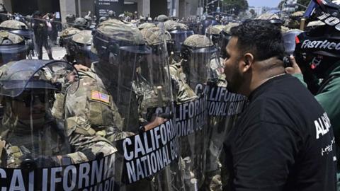 A row of military personnel in camouflage uniforms and helmets stands holding shields as a protester confronts them.