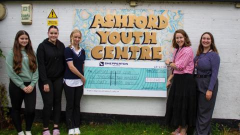 Five women hold a giant cheque from Pinewood to Ashford Youth and Community Club in front of a brick wall which has the youth club's name painted on it.