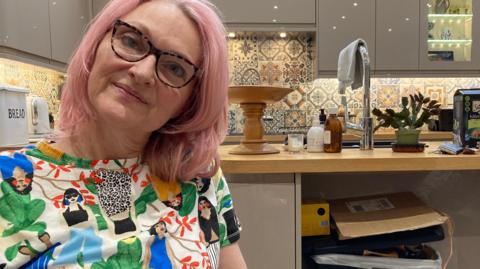 Katherine Ashton, has bobbed pink hair and glasses, is wearing a colourful t-shirt emblazoned with illustrations as she sits in her kitchen