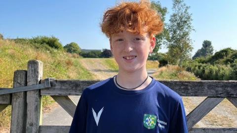 Rowan Thornborough is smiling as he stands in front of a wooden farm gate. He has curly ginger hair and a wears a blue t-shirt with the logo and legend of Cumbria Tennis on the chest..