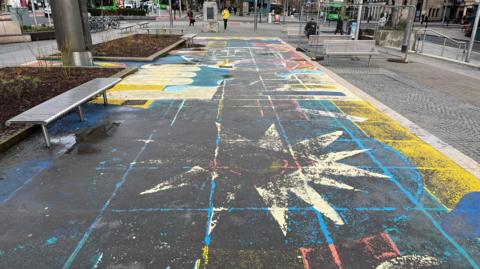 The pavement has been painted with blue, red, yellow and white star-shaped and circular patterns. Black patches are showing through in several places. People are walking in the distance.