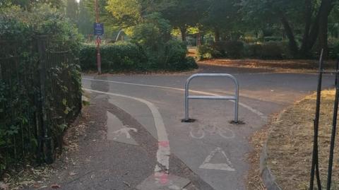 A metal bike barrier is in the middle of a cycle path at the entrance to a park, obstructing cyclist's way in. Around the path are various patches of grass and hedges