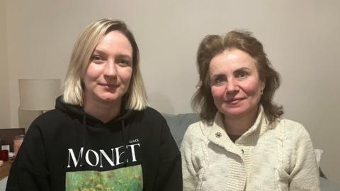 Two women sit side by side indoors on a sofa; one wearing a black hoodie with a Monet print, the other in a light-coloured knitted cardigan. A lamp and small table are visible in the background.