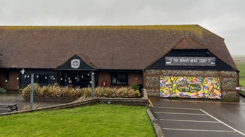 The outside of The Beachy Head Story. It is a long building with black wood on the exterior and a brown tile roof.